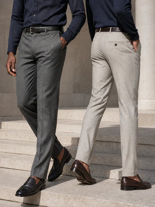 Two men wearing slim-fit dress pants, one in charcoal gray and the other in light beige, paired with navy shirts and brown loafers, walking down stone steps.