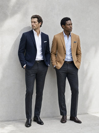 Two men standing outdoors against a light gray wall, each wearing a tailored blazer and dark trousers. The man on the left wears a navy blazer with a white shirt, and the man on the right wears a tan blazer with a light blue shirt.