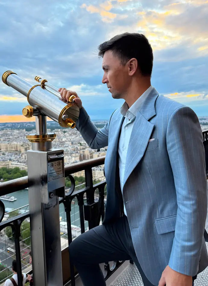 Man in a light blue suit using a telescope with a cityscape and river in the background