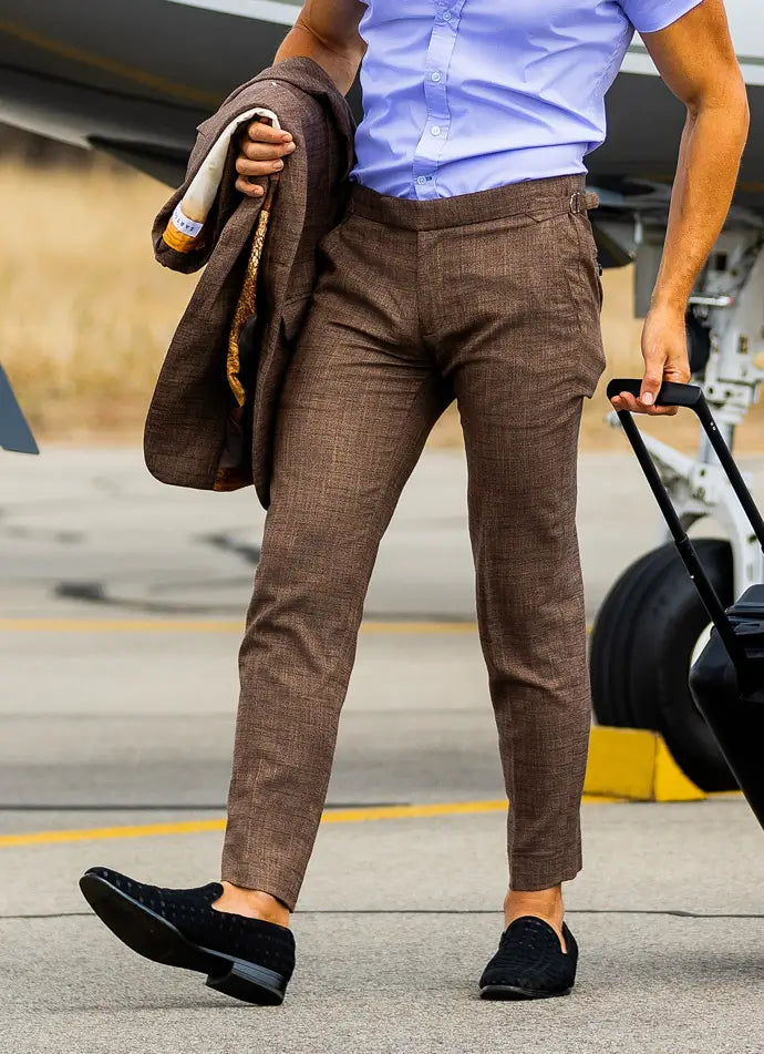 Person wearing brown pants and a blue shirt, holding a jacket and pulling a suitcase on a tarmac