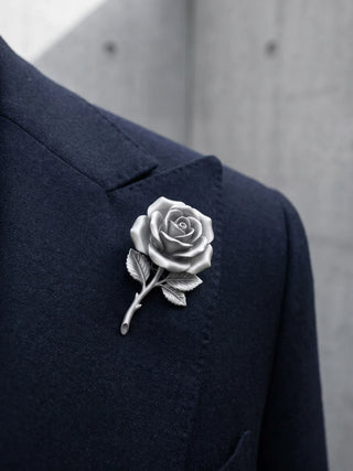 A detailed silver rose boutonniere brooch pinned to the lapel of a dark navy blue suit jacket, set against a blurred gray concrete wall background.