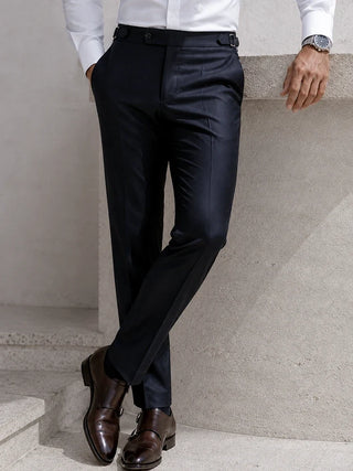 A man wearing slim-fit black trousers, a white dress shirt, and dark brown leather dress shoes, standing against a light gray concrete wall.