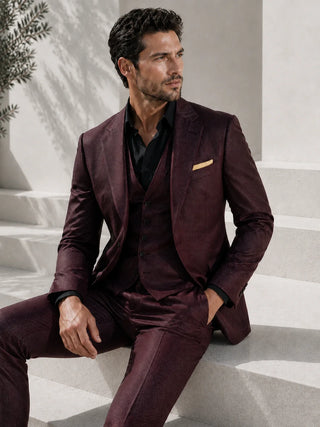 A man wearing a three-piece burgundy suit with a black shirt and a gold pocket square, sitting on light-colored stone steps.