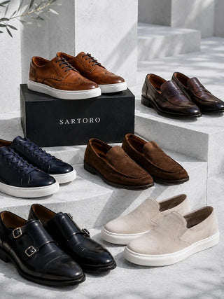 A collection of Sartoro shoes arranged on light gray stone steps, including brown oxford sneakers, dark brown penny loafers, navy low-top sneakers, brown suede loafers, black double-strap loafers, and beige slip-on loafers