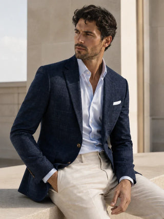 A man wearing a navy blue textured blazer, a white dress shirt, and beige trousers, sitting on a stone ledge in an outdoor architectural setting.