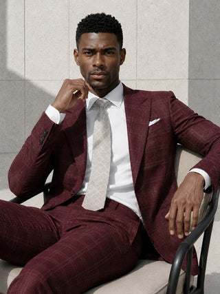 A man wearing a burgundy windowpane plaid suit with a white dress shirt and a light beige patterned tie, sitting on a chair against a light gray tiled wall.