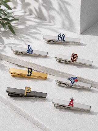 Six silver-toned baseball and sports clip hair ties with team logos, arranged on a light textured surface with soft shadows and a small plant leaf in the corner.