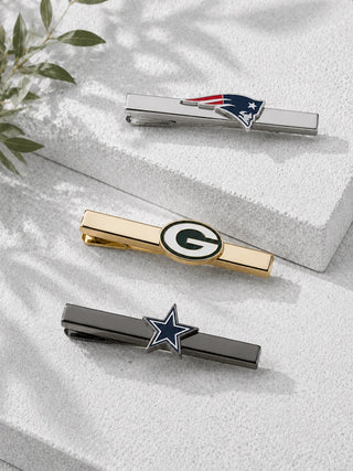 Three silver-tone tie bars with sports team logos, including the New England Patriots, Green Bay Packers, and Dallas Cowboys, arranged on a textured white block with soft leaf shadows.