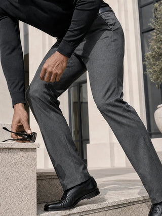 A man wearing slim-fit charcoal gray dress pants, a black long-sleeve shirt, and black leather dress shoes, leaning over stone steps while holding a pair of sunglasses.