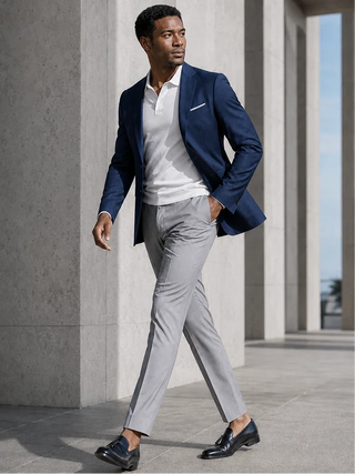 A man walking outdoors in a stylish outfit consisting of a navy blue blazer, white polo shirt, light gray trousers, and black loafers, set against a modern concrete architectural background.