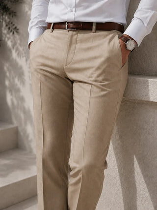A person wearing beige chinos with a brown leather belt, styled with a white dress shirt and a brown leather watch, leaning against a light stone wall.
