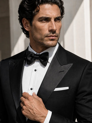 A man wearing a classic black tuxedo with satin lapels, a white pinstripe dress shirt, a black bow tie, and a white pocket square, set against a soft-focus architectural background.