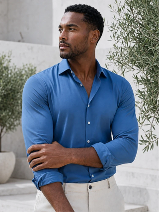A man wearing a long-sleeve blue button-down shirt and light beige trousers, standing outdoors against a light stone wall with green plants.