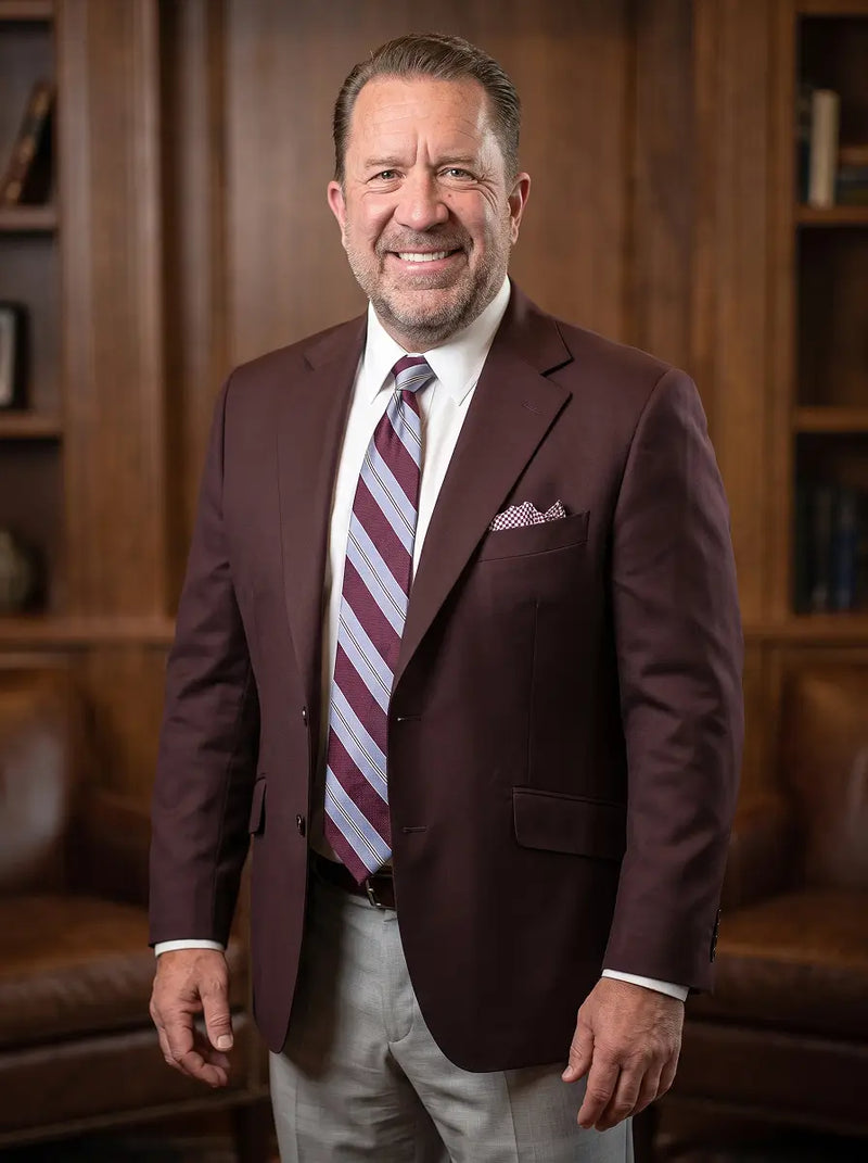 A smiling man in a maroon blazer, white dress shirt, and a striped burgundy and silver tie, standing in front of a warm wood-paneled wall and leather chairs.