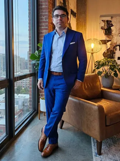A man wearing a bright blue two-piece suit, light blue dress shirt, and brown leather dress shoes stands in a modern room with a large window, brick wall, and brown leather armchair.