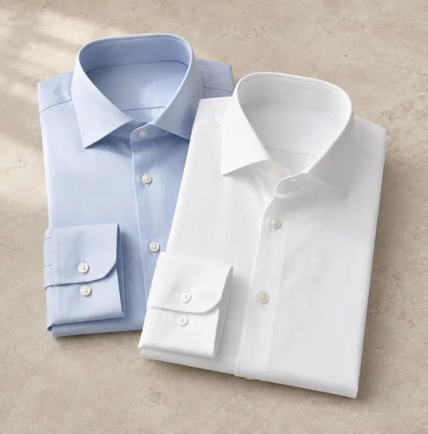 Two folded dress shirts on a light beige surface: one light blue shirt with a spread collar and button-down cuffs, and one white shirt with a pointed collar and button-down cuffs.