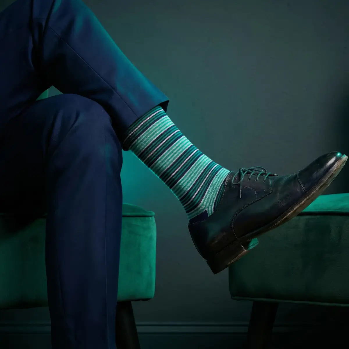 Person wearing blue striped socks and dark shoes sitting on a green stool against a dark background990