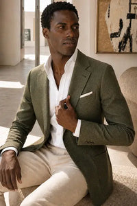 A man wearing a textured olive green blazer over a white dress shirt and beige trousers, sitting in a modern indoor setting.