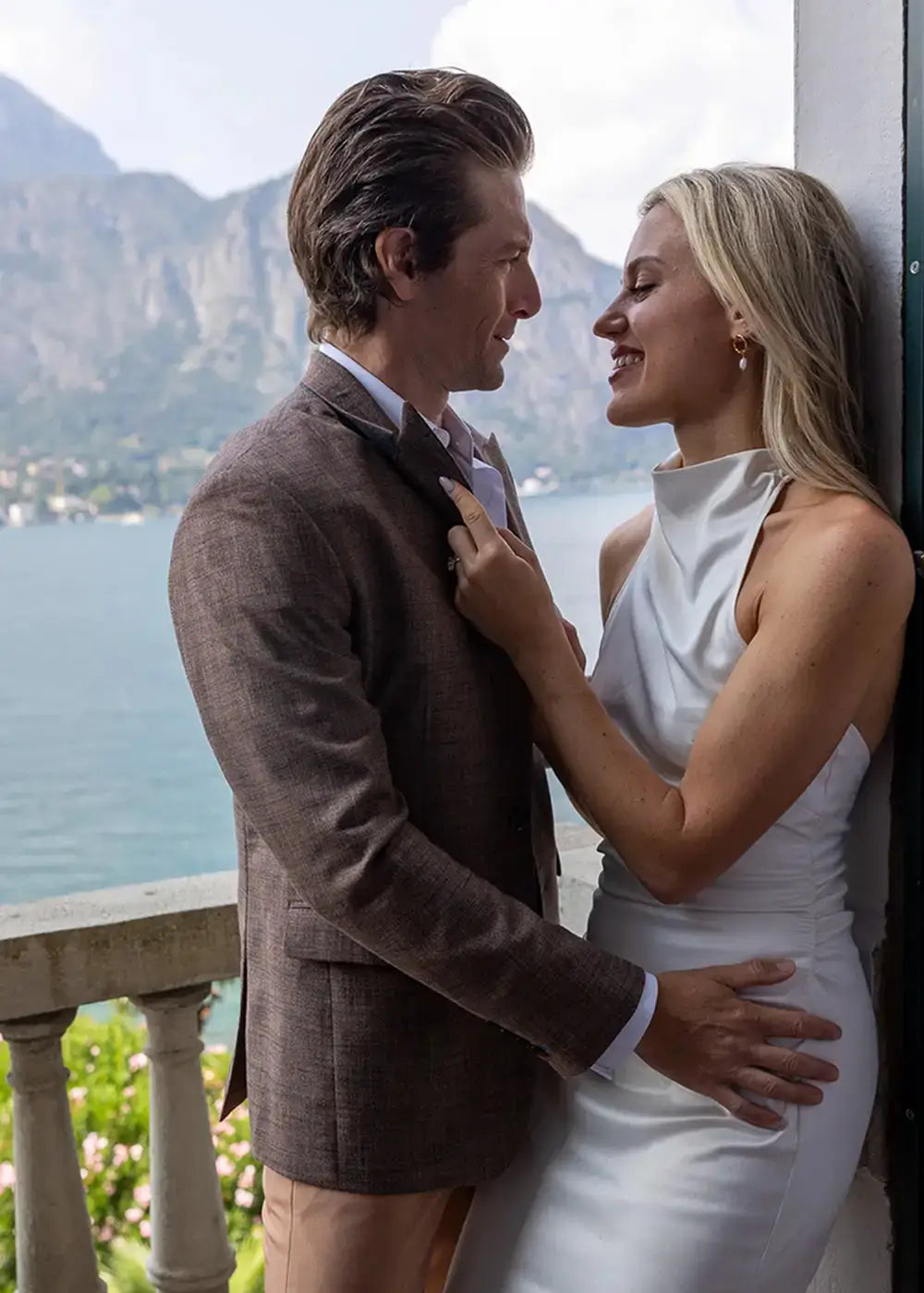 A man in a brown textured blazer and a woman in a white sleeveless dress embracing on a balcony overlooking a scenic lake and mountains.