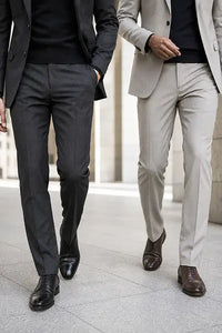 Two men walking outdoors wearing slim-fit dress pants, one in dark charcoal gray and the other in light beige, both styled with black shirts and brown leather dress shoes.