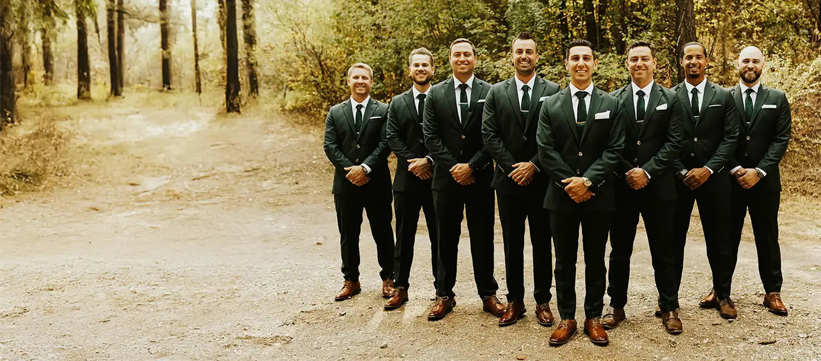Seven men in matching dark green suits and ties standing together on a dirt path in a wooded outdoor setting.