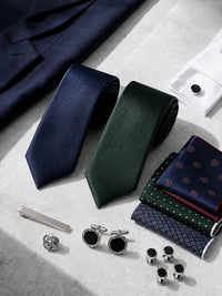 A styled flat-lay of men's formal accessories on a light beige stone surface, including two rolled neckties in navy and forest green, folded patterned pocket squares, silver cufflinks, cufflink buttons, a tie bar, and a pocket square holder.