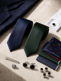 A styled flat-lay of men's formal accessories on a light beige stone surface, including two rolled neckties in navy and forest green, folded patterned pocket squares, silver cufflinks, cufflink buttons, a tie bar, and a pocket square holder.