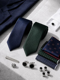 A styled flat-lay of men's formal accessories on a light stone surface, including two rolled neckties in navy and forest green, folded patterned pocket squares, silver cufflinks, cufflink buttons, a tie bar, and a pocket square, with part of a navy suit jacket and a white shirt cuff visible.