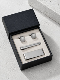 A men's formal accessory set including silver cufflinks, a tie clip, and a tie bar, presented in an open black textured gift box on a light gray stone surface.