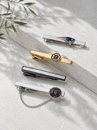 Four men's tie clips arranged diagonally on a textured white surface with soft leaf shadows. The clips include silver, gold, and dark gray metallic finishes, with team emblems including the New England Patriots, Boston Bruins, and New York Rangers.