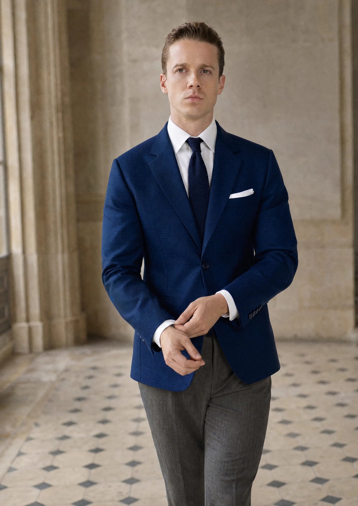 Man in a royal blue microcheck jacket, white shirt and navy tie walking through a limestone townhouse hall892