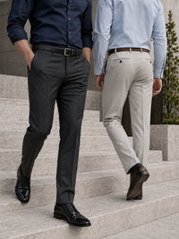 Two men walking down outdoor stone steps, shown from the waist down. One wears dark charcoal gray dress pants with a black belt and black dress shoes, and the other wears light beige dress pants with a brown belt and brown dress shoes.