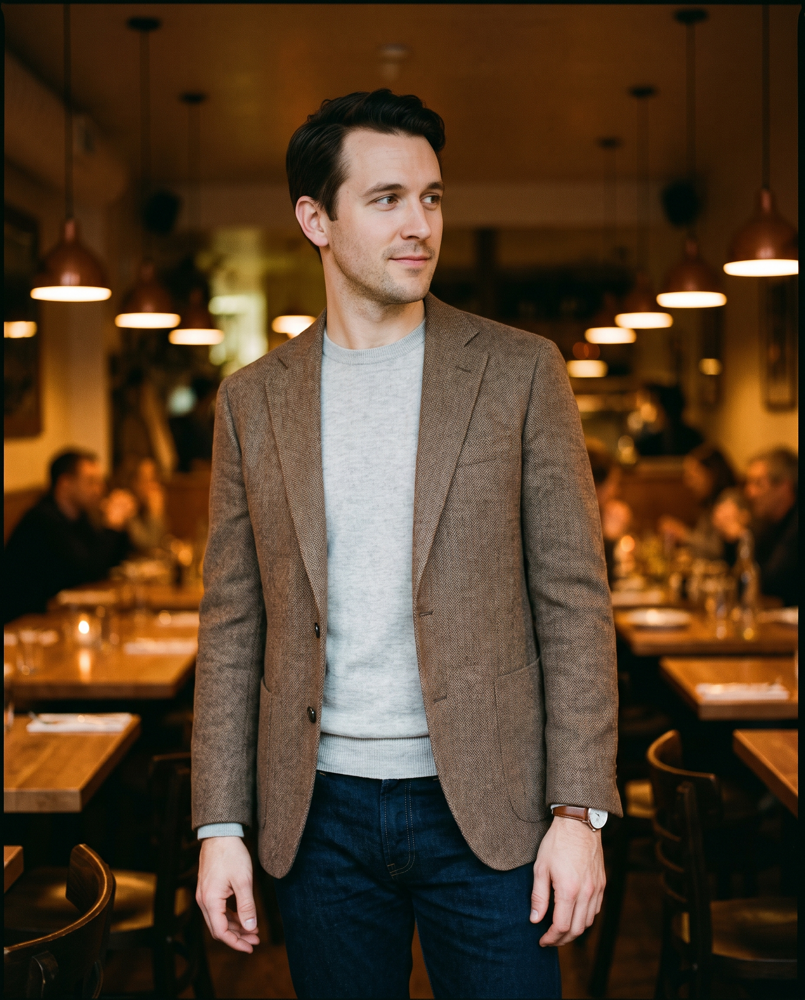 A man in a herringbone sport coat, dark jeans, and camel suede loafer standing in a warmly lit restaurant interior — a practical example of smart casual in a social setting