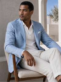 A man seated in a chair wearing a light blue blazer over a white polo shirt and white trousers, photographed in a bright architectural interior.