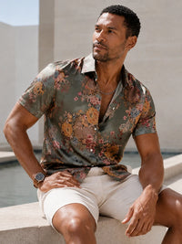 A man wearing a short-sleeve button-up shirt with a muted olive green base and a warm-toned floral print, paired with light beige shorts, sitting outdoors.