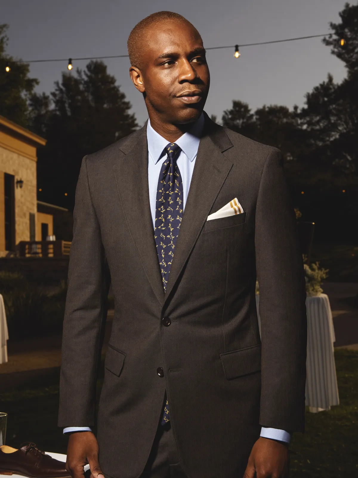 Wedding guest in a charcoal cocktail suit with pale blue shirt and patterned silk tie at an outdoor wedding cocktail hour in soft early-evening light