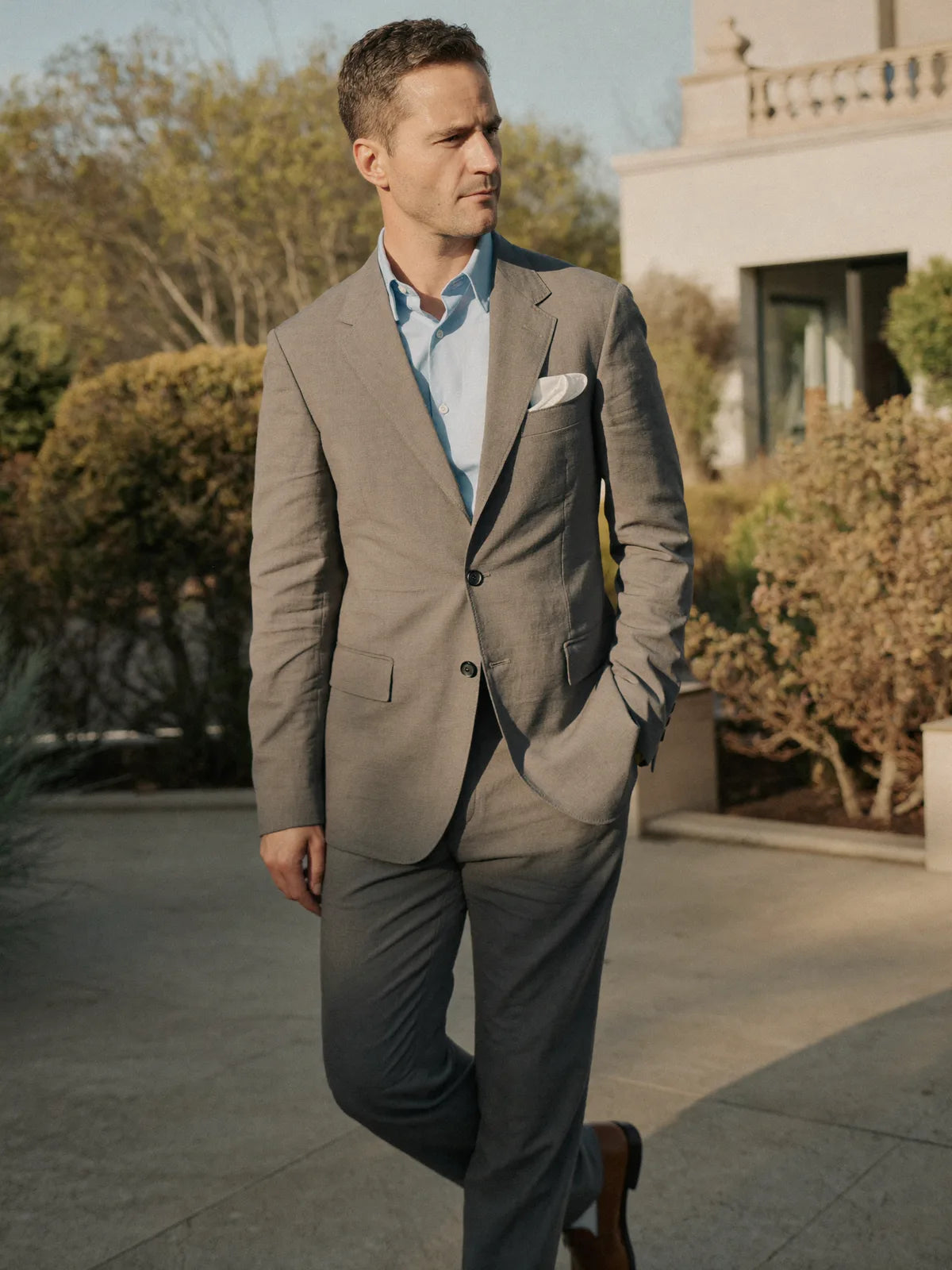 Man in a mid-gray daytime cocktail suit with open-collar pale blue shirt and brown loafers in a sunlit garden gallery setting