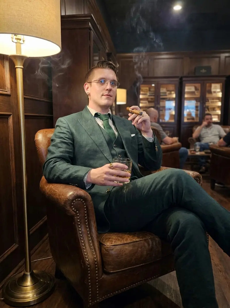A man in a dark green suit and green tie sits in a brown leather armchair inside a dimly lit wood-paneled lounge, holding a cigar and a glass of whiskey.