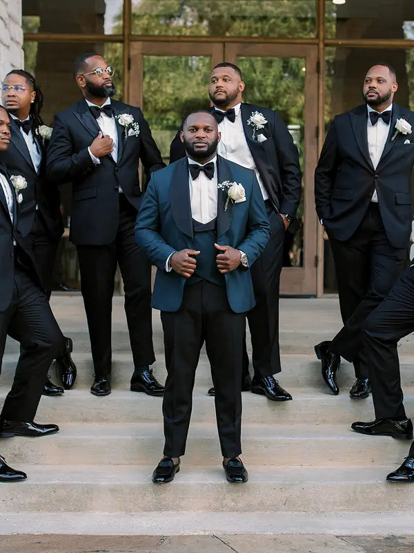 A group of men in formal evening wear standing on outdoor stone steps. The central figure wears a dark teal tuxedo jacket with black trousers, a black bow tie, a white dress shirt, and a white rose boutonniere.