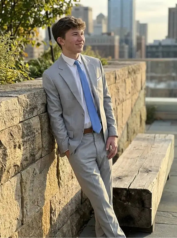 A young man wearing a light gray suit, white dress shirt, and light blue tie leans against a stone wall outdoors with a city skyline in the background.