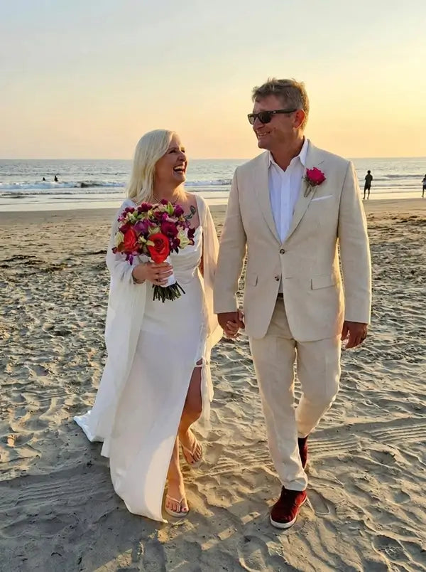 A bride in a white wedding dress and a groom in a light beige suit walk hand-in-hand on a sandy beach at sunset. The bride holds a vibrant multicolor bouquet.