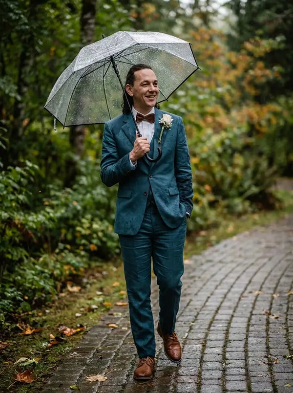 A man walking on a wet cobblestone path in a forest, holding a clear umbrella. He is wearing a teal blue three-piece suit, a white dress shirt, a brown bow tie, and brown leather shoes.