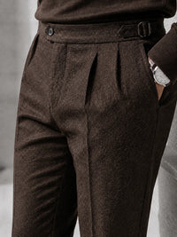 Close-up of a person wearing dark brown pleated wool trousers with a matching belt, shown from the waist down against a neutral gray background.