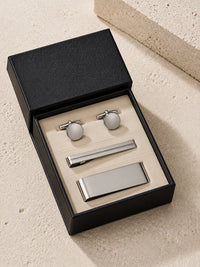 A men's formal accessory set in a black presentation box, including silver-toned cufflinks with white oval inlays, a silver tie clip, and a silver toner bar, displayed on a beige textured surface.