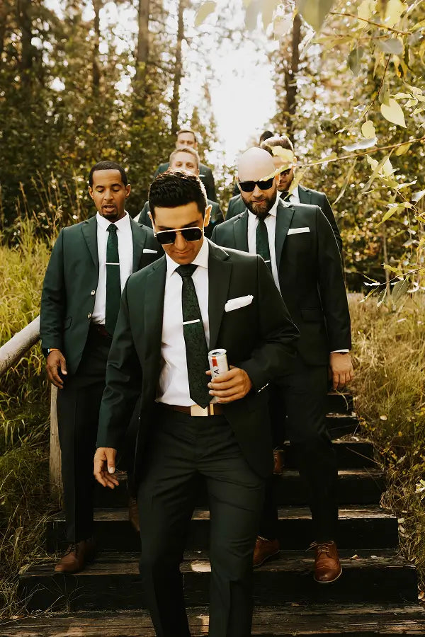 A group of men in matching dark green suits, white shirts, and black ties walk down outdoor wooden steps in a wooded setting. The front man wears sunglasses and holds a drink can.