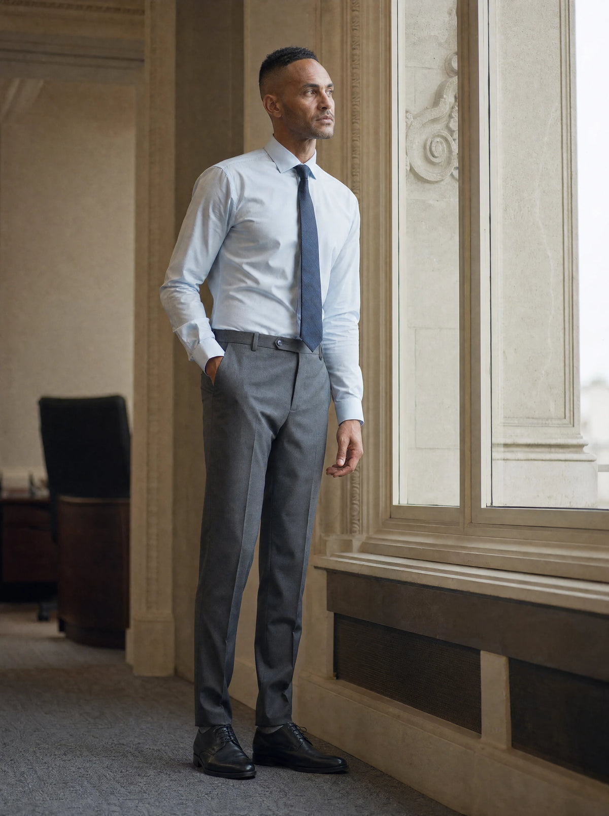 Man in grey charcoal crosshatch trousers and pale blue shirt standing at an office window520