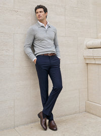 A man wearing a light gray long-sleeve polo sweater, navy blue dress pants, a brown belt, and brown leather dress shoes, leaning against a beige stone wall.