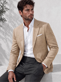 A man wearing a beige textured blazer over a white dress shirt and dark trousers, seated against a light gray wall with soft shadows.