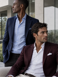 Two men in tailored suits posing outdoors. The seated man in the foreground wears a burgundy blazer over a white shirt, and the standing man behind wears a navy blazer over a light blue shirt.