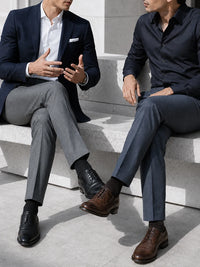 Two men in tailored suits sitting on white stone steps outdoors, wearing navy and gray blazers with brown and black dress shoes.